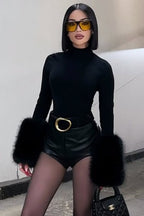 Person wearing a black turtleneck, black leather shorts with fur trim, and yellow sunglasses against a plain background.