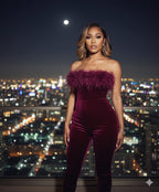 Woman in a burgundy strapless jumpsuit with feather details against a city skyline at night.