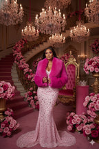 Woman in a pink dress and fur coat standing in an opulent room with chandeliers and floral arrangements.